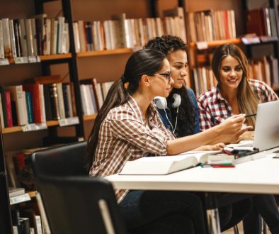 Three young women collaborate on a project in a library, utilizing a shared workspace. This highlights the convenience of reserving study areas with a space booking app for efficient teamwork and learning.