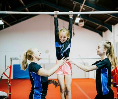 AdobeStock_244454934 Two coaches assist a young gymnast practicing on the high bar in a brightly lit gym. The scene highlights the importance of safety, teamwork, and structured training programs, making it ideal for gymnastics management software that streamlines scheduling and class organization