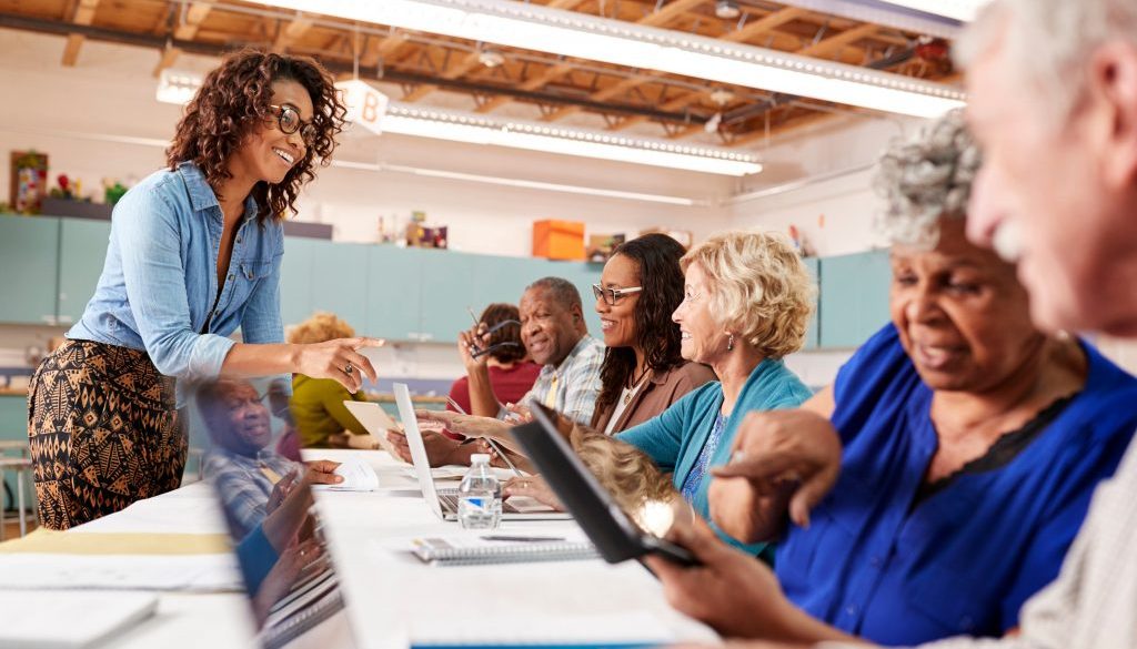 A group of older adults engages in a technology class at a community center, using laptops and tablets while a friendly instructor provides guidance. This scene highlights how community center software can streamline class scheduling, making it easier for centers to offer impactful learning experiences.