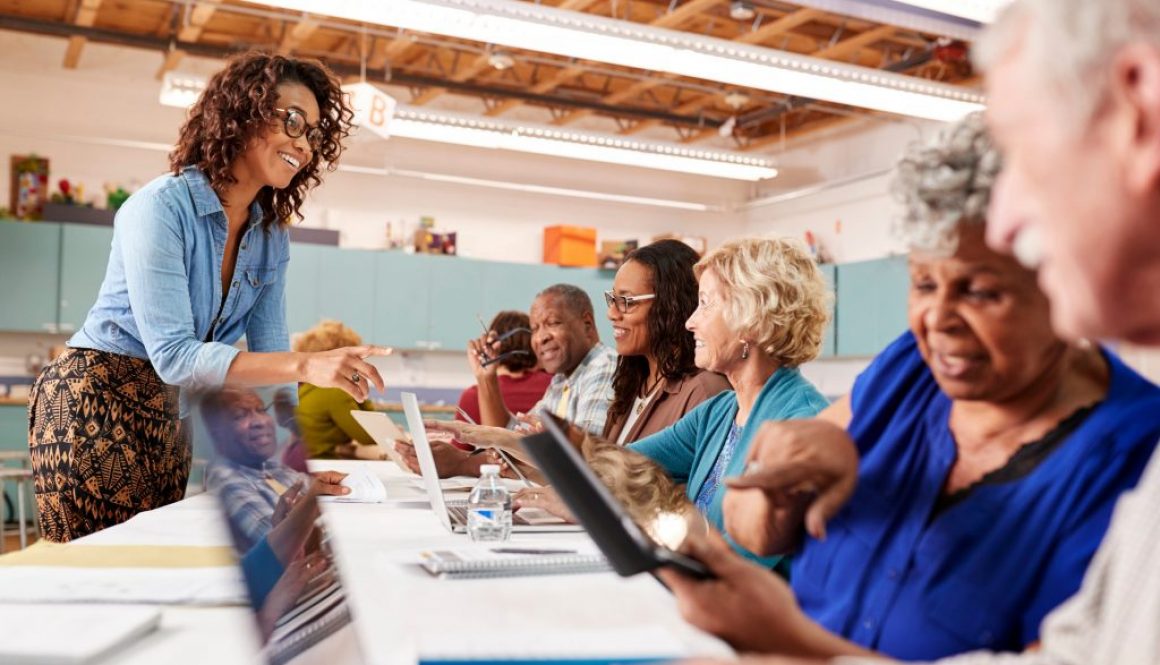 A group of older adults engages in a technology class at a community center, using laptops and tablets while a friendly instructor provides guidance. This scene highlights how community center software can streamline class scheduling, making it easier for centers to offer impactful learning experiences.