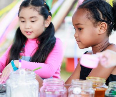Lovely Asian and African girls playing dough together at playground, summer camp learning Two young girls engaged in a creative activity with jars of colorful materials at a summer camp, highlighting the fun and variety of programs that can be organized and managed efficiently with summer camp registration software.