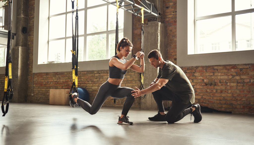 Personal trainer guiding a woman during TRX squats at the gym, with sessions scheduled using athletic training software.
