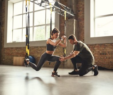 Doing squat exercise. Confident young personal trainer is showing slim athletic woman how to do squats with Trx fitness straps while training at gym. Personal trainer guiding a woman during TRX squats at the gym, with sessions scheduled using athletic training software.