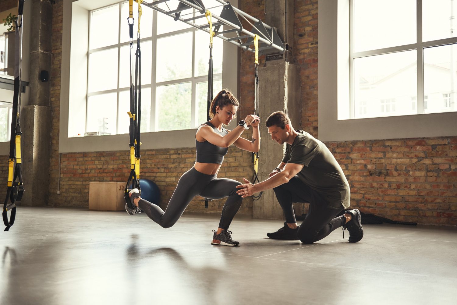 Doing squat exercise. Confident young personal trainer is showing slim athletic woman how to do squats with Trx fitness straps while training at gym. Personal trainer guiding a woman during TRX squats at the gym, with sessions scheduled using athletic training software.