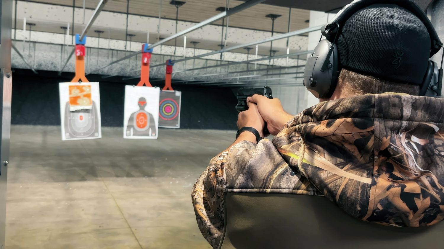 Shooting Practice at Indoor Gun Range. A person wearing camouflage and protective gear takes aim at paper targets during shooting practice at an indoor gun range.